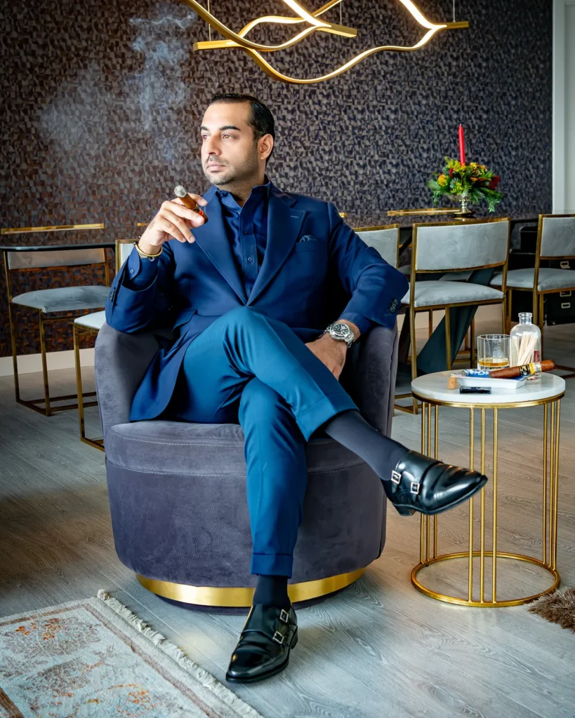 Retrato de Kabir Sukhwani sentado en un sillón de su casa con traje azul oscuro y un puro.